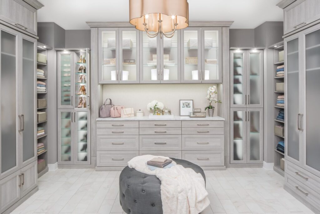 Bright walk-in closet with gray wood cabinets, glass-front uppers, and a central dresser; a plush ottoman sits in the middle.