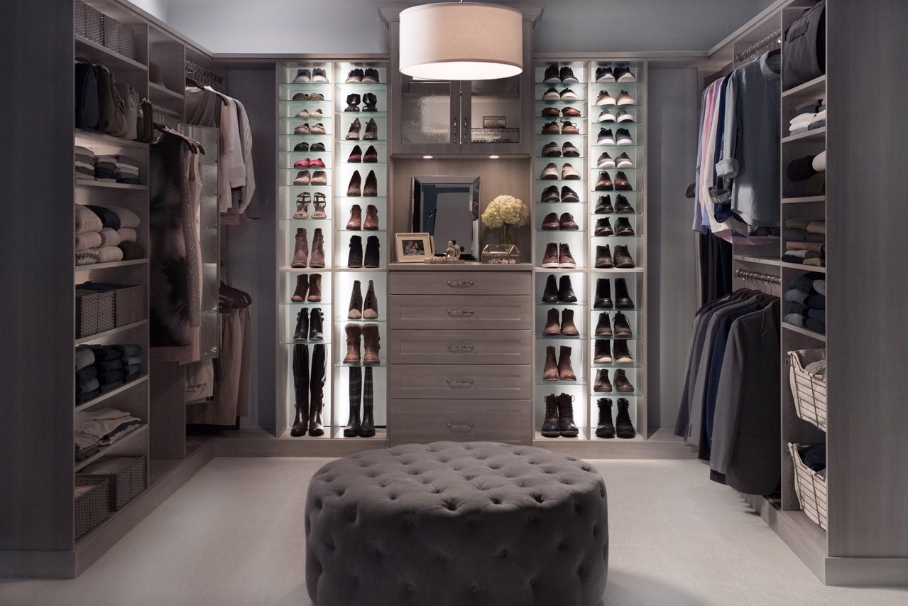 D 291 1 Space Crafters Co Walk-in closet with illuminated shoe shelves surrounding a central dresser, framed photos, and a plush tufted ottoman in the center.